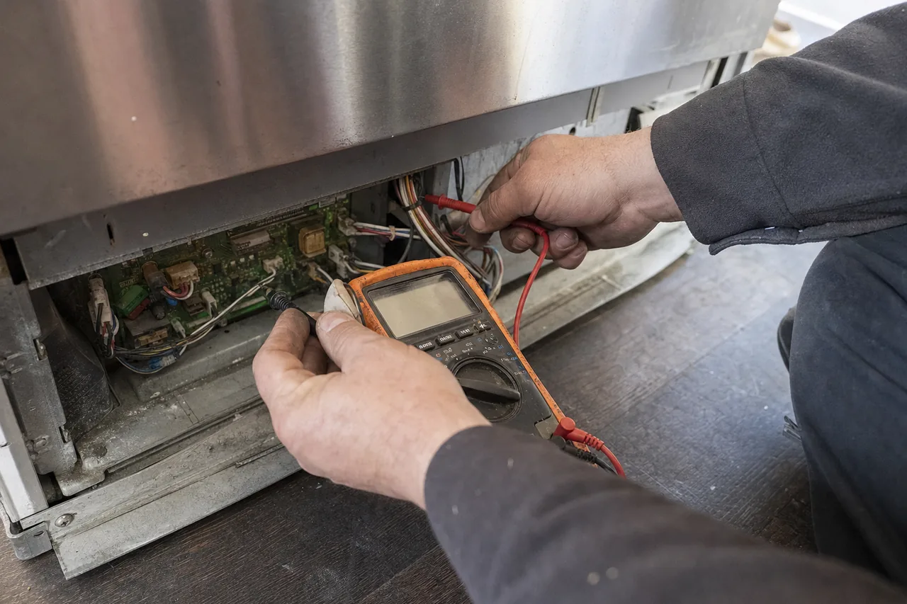 A technician kneeling on a wooden floor with a multimeter in one hand, red and black probes touching a control board that has been slid out from beneath a stainless cabinet, wiring loom and connectors exposed.
