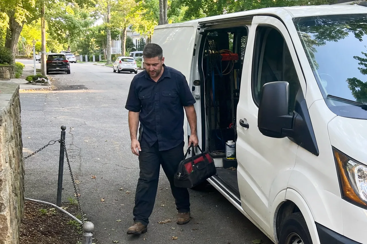 A technician stepping away from the open side door of a plain white van parked on a shaded residential street, a soft tool bag in one hand and hose reels and racking visible inside the van.