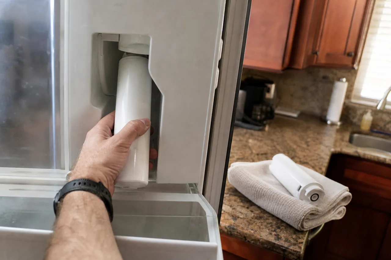A hand seats a white filter cartridge in its housing inside an open refrigerator, with a second cartridge resting on a folded towel on the granite counter by the cherry cabinets